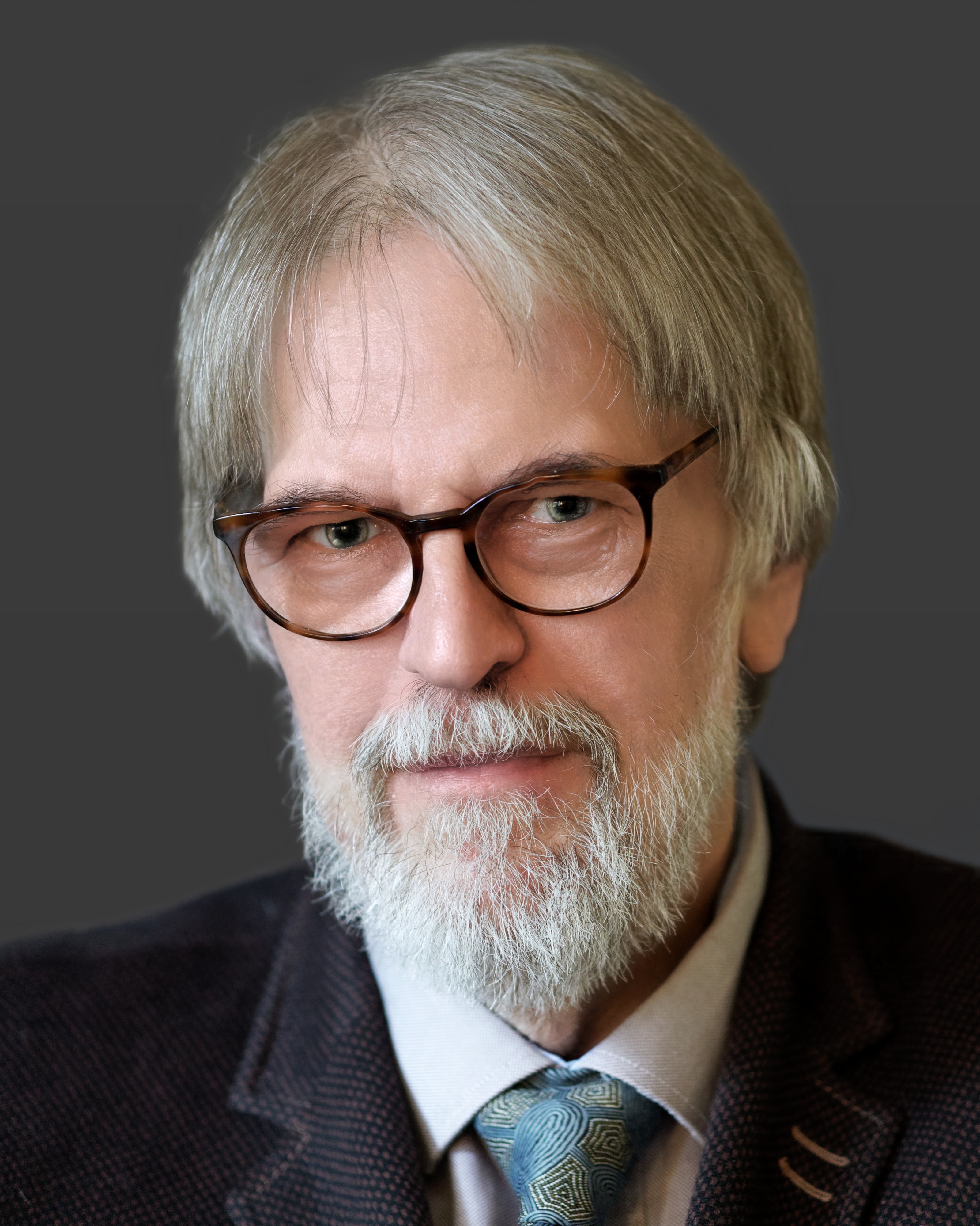 An older man with gray hair, glasses, and a beard wears a suit jacket, light shirt, and patterned tie, posing against a neutral background.