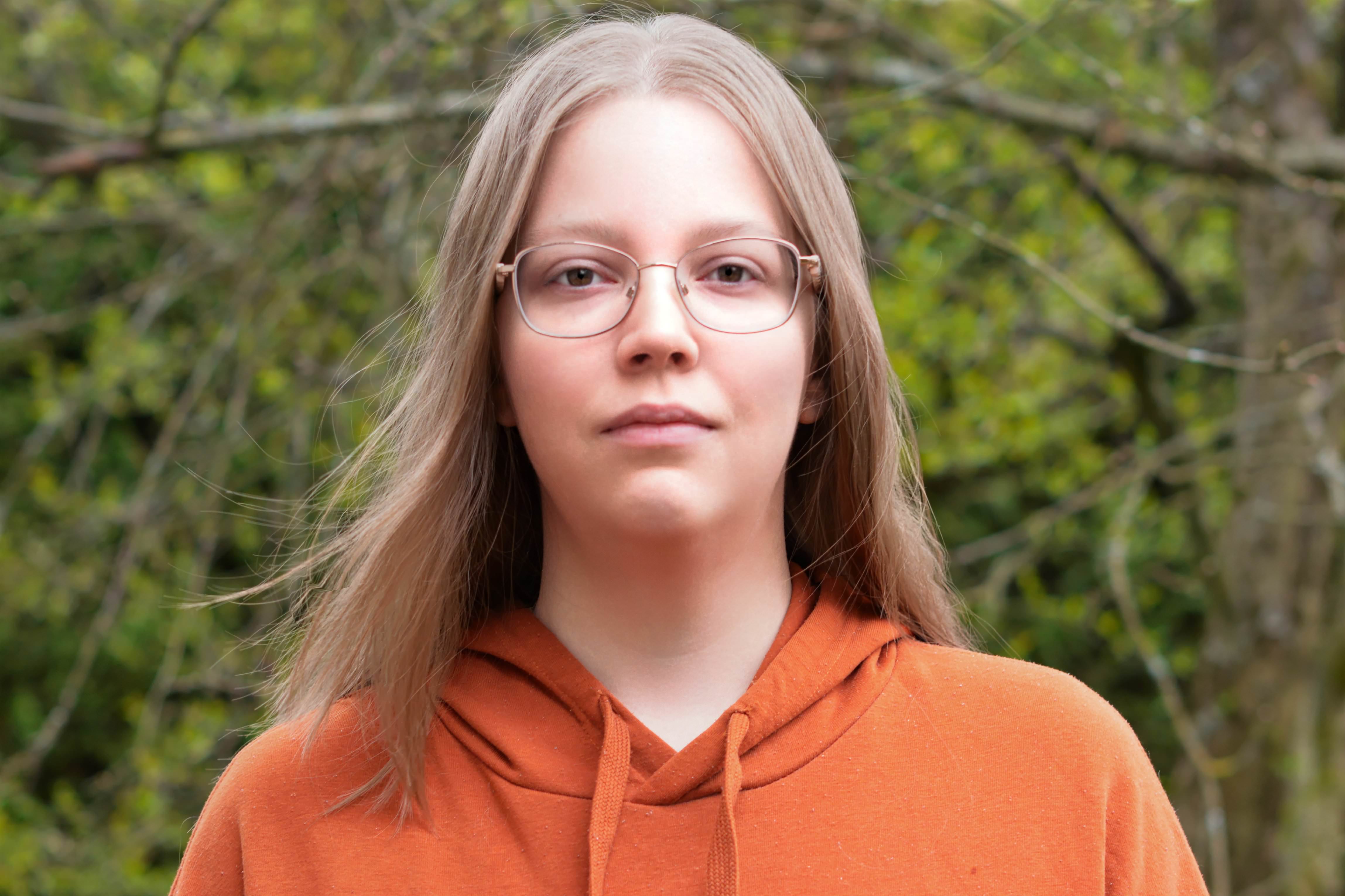 A young woman with straight, light brown hair and glasses wears an orange hoodie, 
          standing outdoors in front of green, leafy trees.