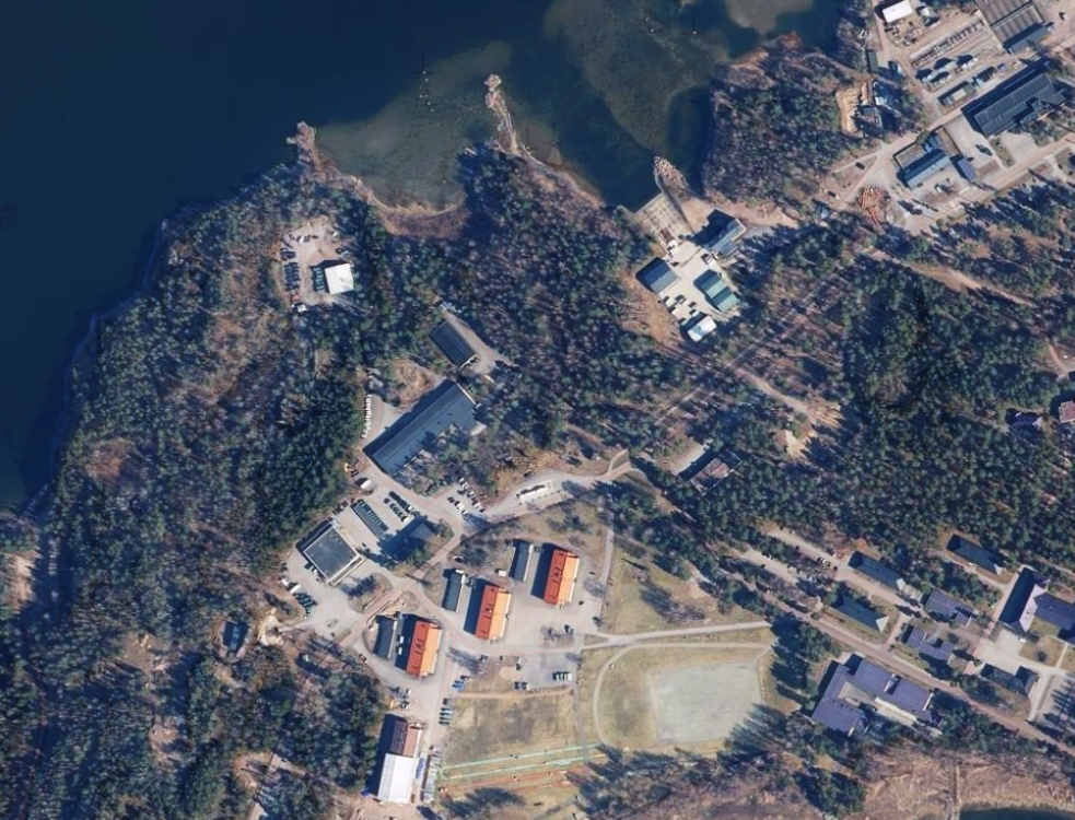 Aerial view of a wooded area with scattered buildings, parking lots, and a body of water at the top left.