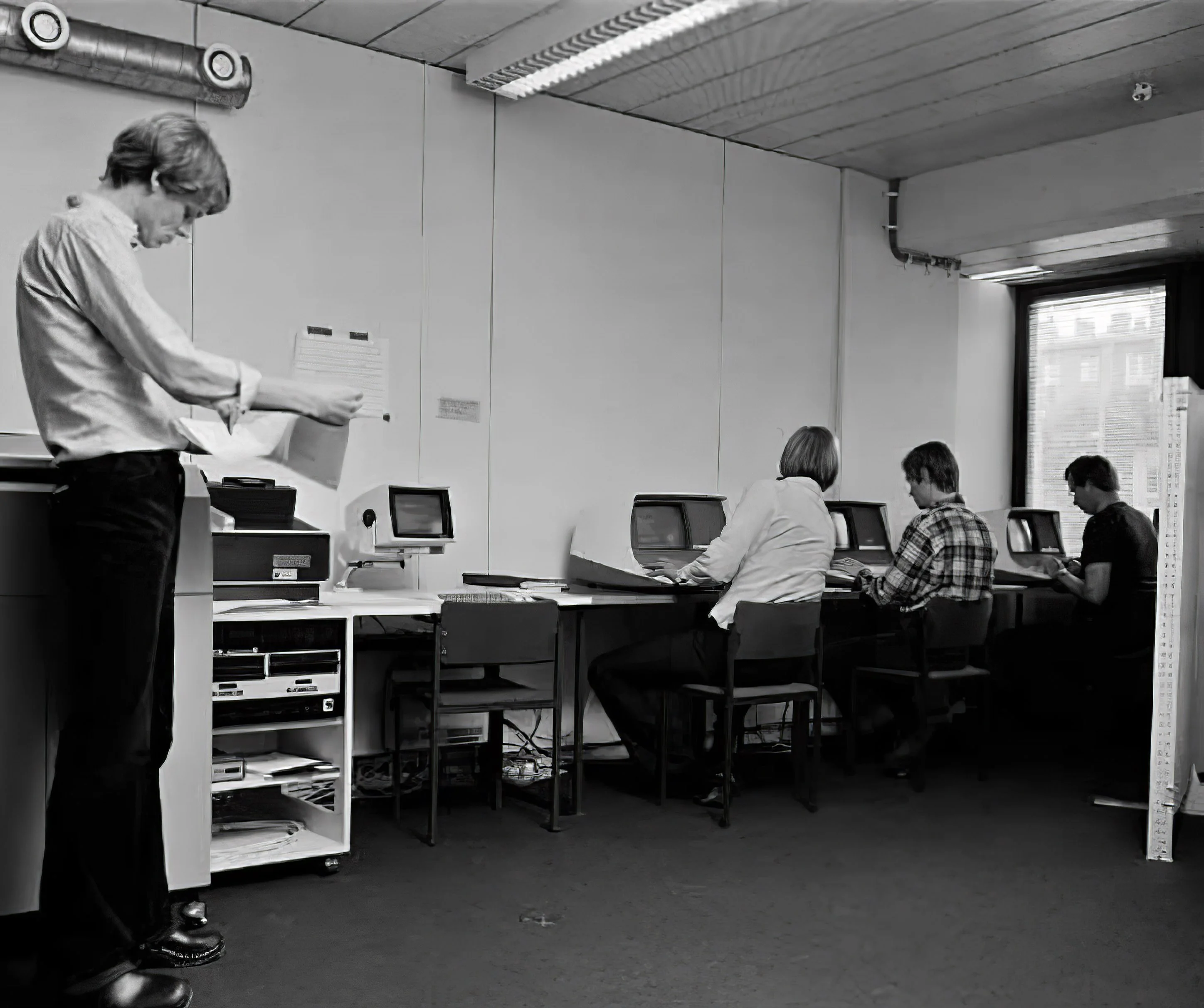 A man stands and examines a printout and three men sit in front of display terminals.