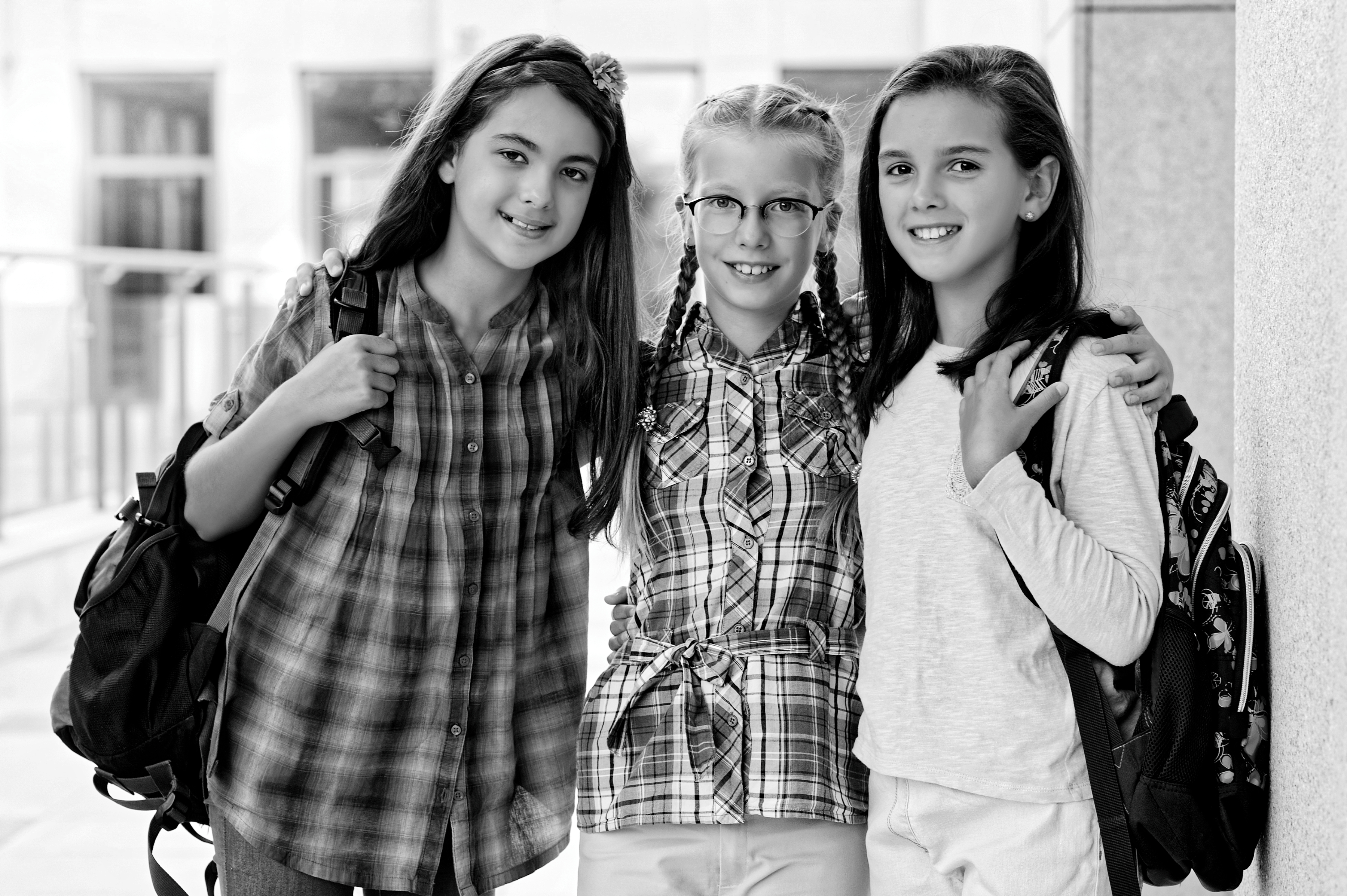 Black-and-white photo of three smiling schoolgirls with backpacks, standing arm in arm in a school hallway, conveying friendship and confidence.